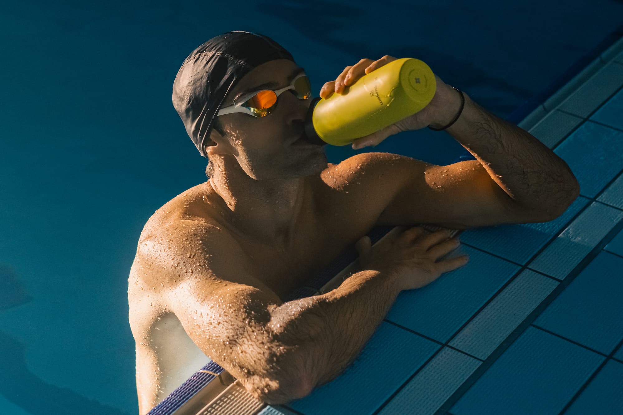 A swimmer wearing goggles and a swim cap resting at the pool edge, drinking from a yellow waterdrop bottle.