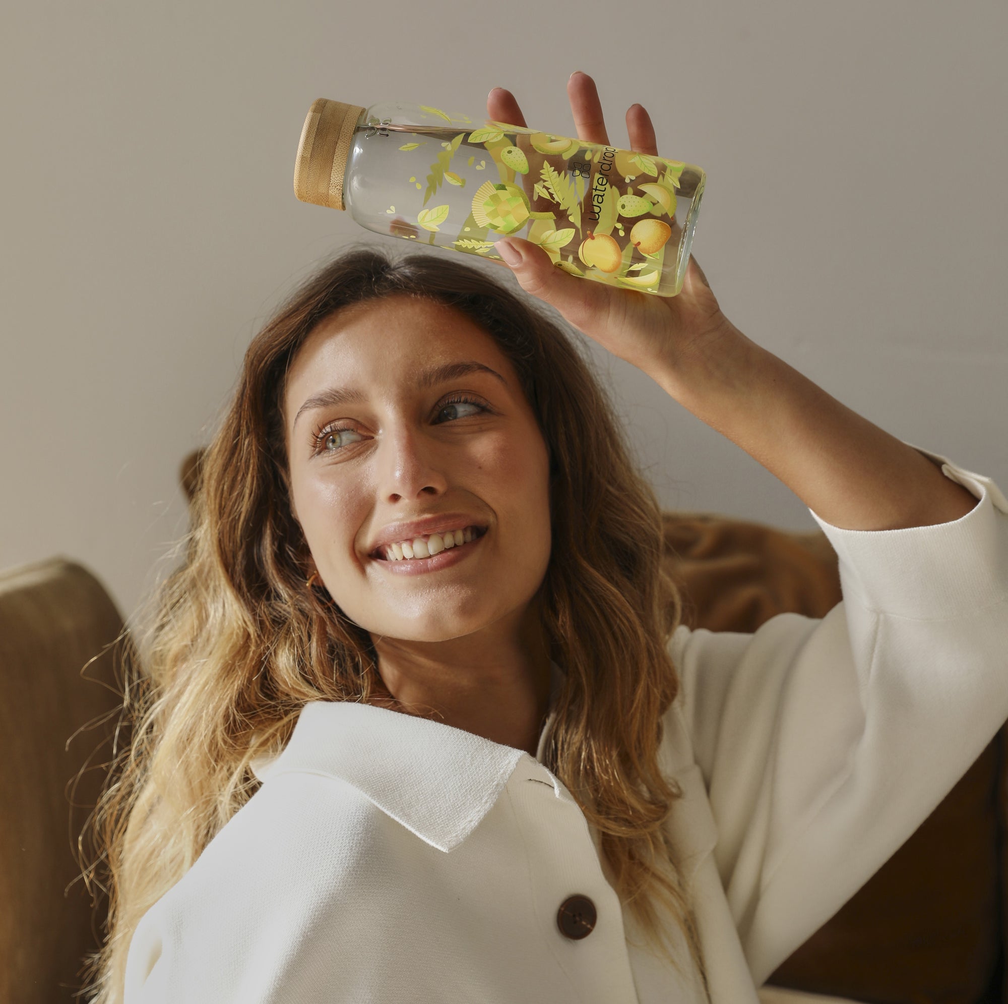 A smiling woman holding a decorative waterdrop glass bottle with vibrant yellow floral and fruit illustrations above her head.