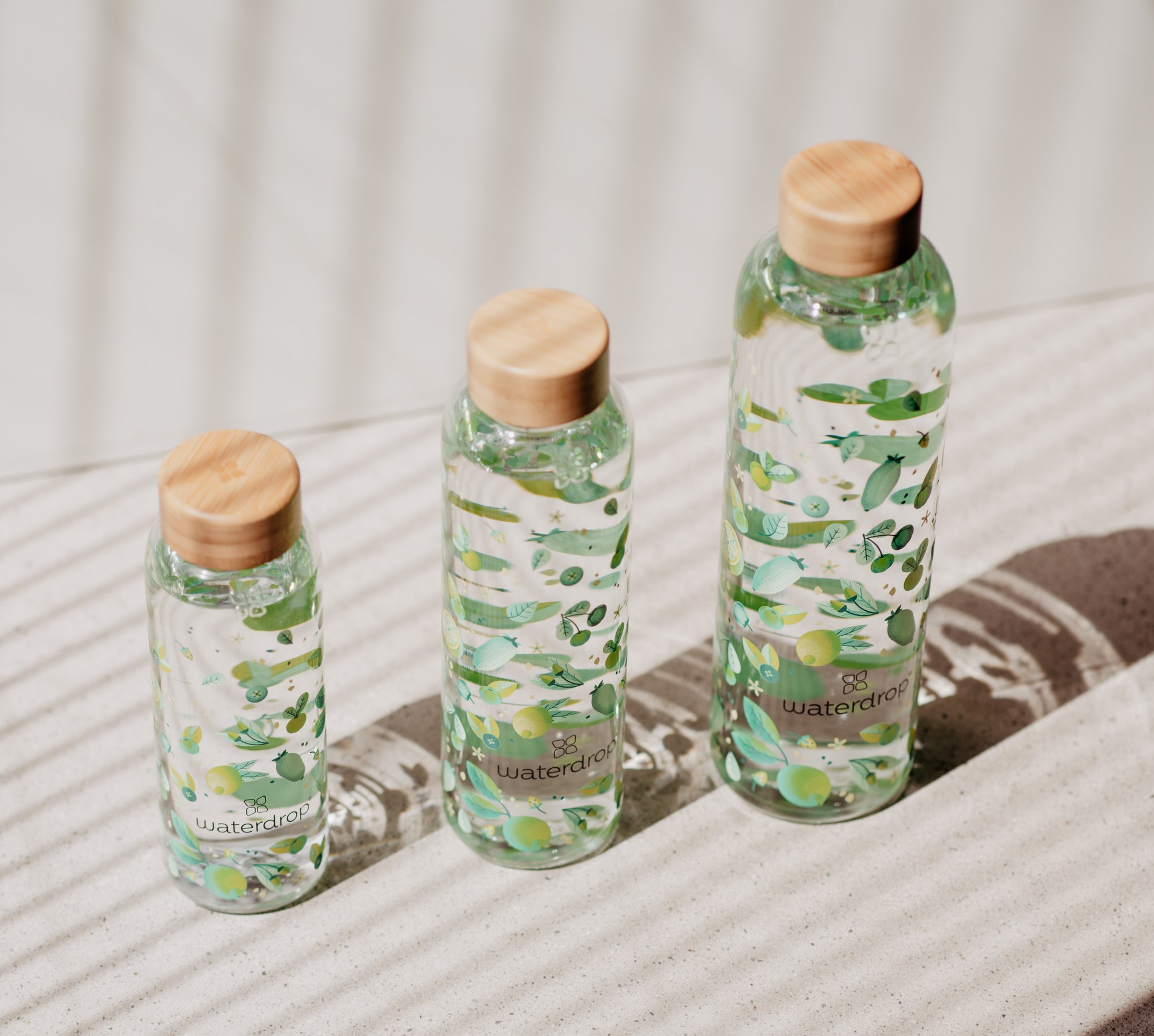 Three waterdrop edition glass bottles of varying sizes with green botanical designs, arranged on a textured surface and casting soft shadows.