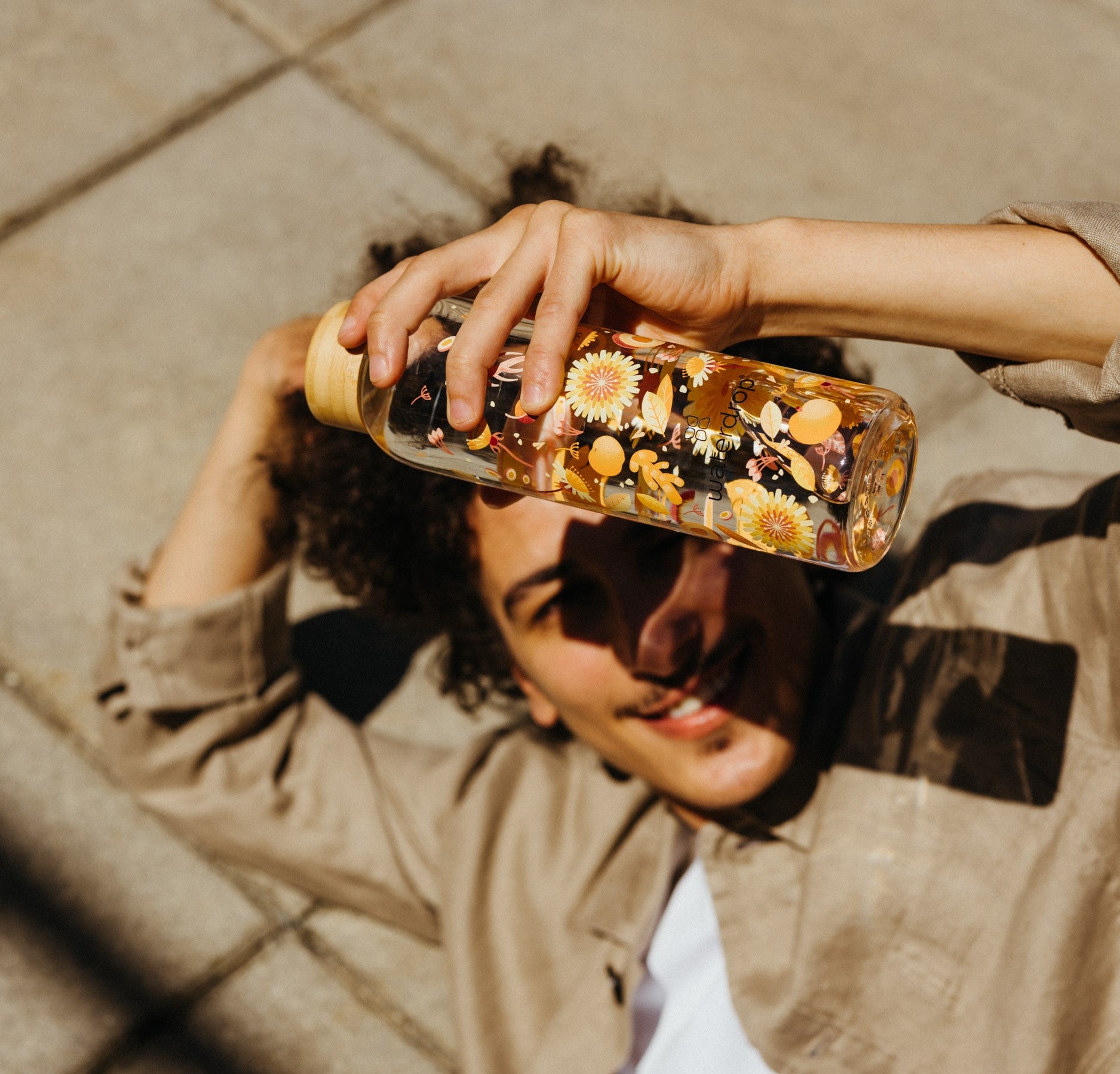 A man lying on the ground, smiling and holding a decorative waterdrop glass bottle with floral and fruit illustrations above his head, casting a shadow on his face.