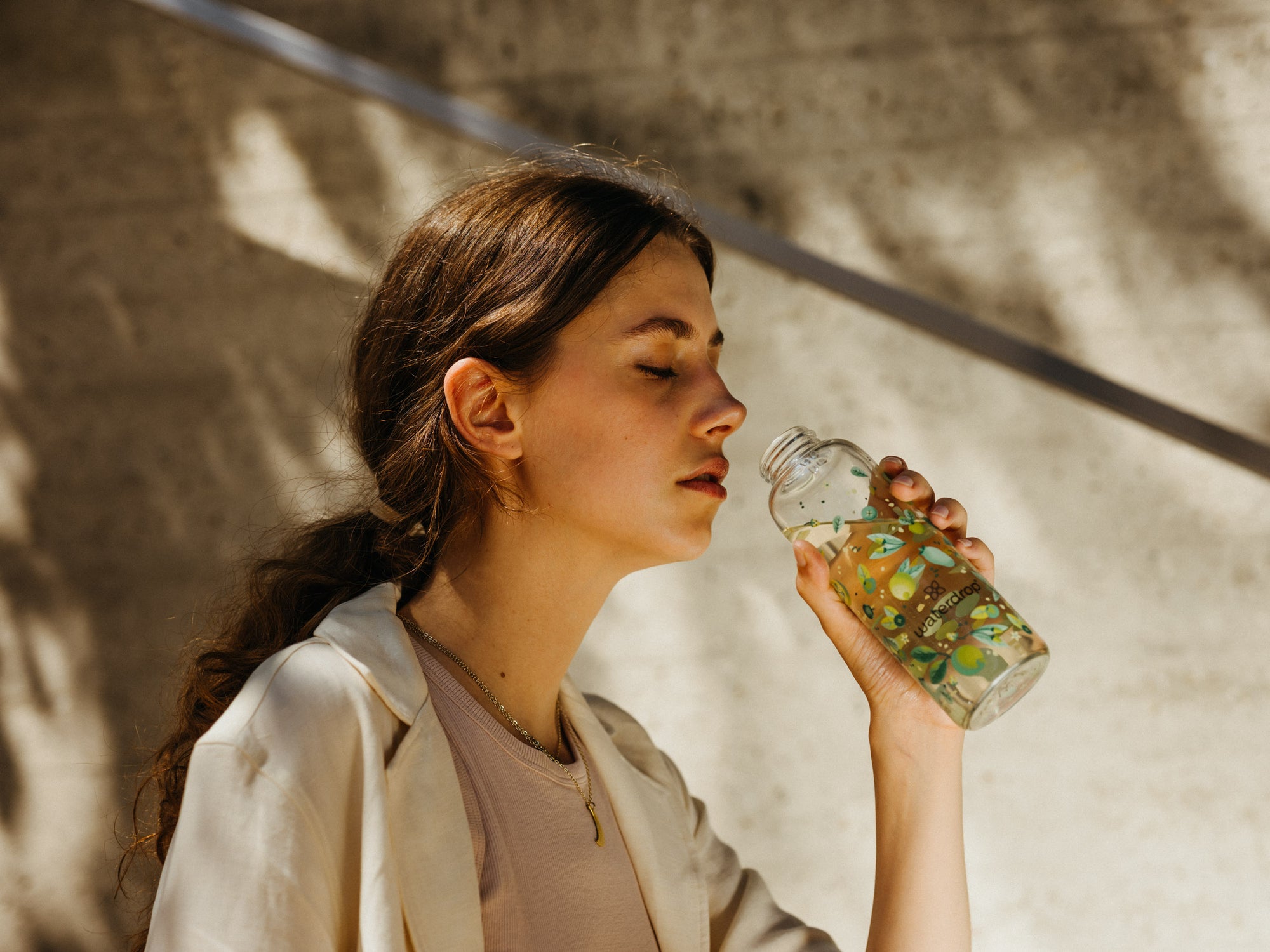 A woman with closed eyes holding a decorative waterdrop bottle with green and yellow designs, preparing to take a sip in a sunlit outdoor setting.