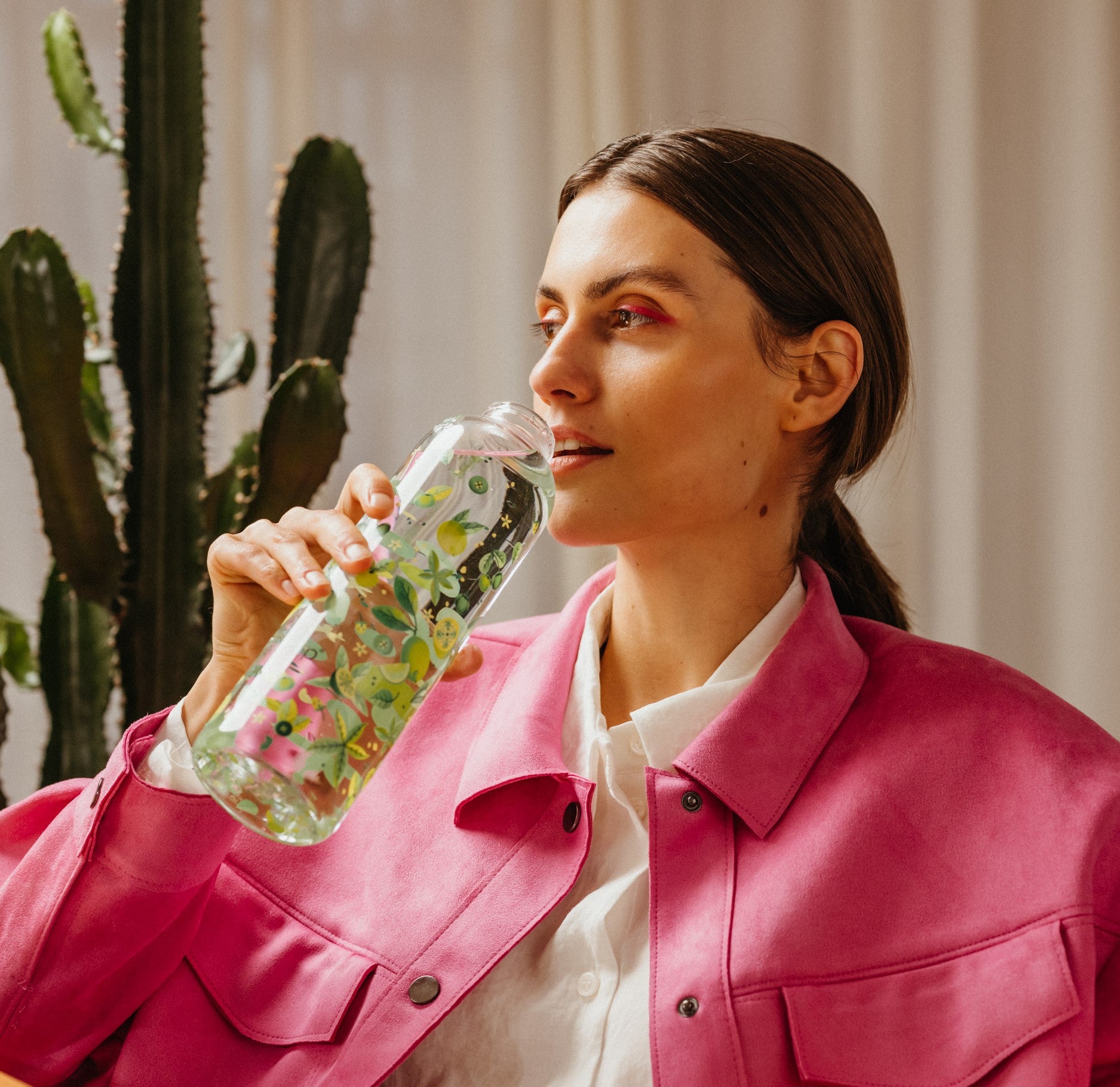 Stylish woman in a pink jacket drinking from a waterdrop bottle with a floral design. Promoting hydration and wellness with a fashionable touch, featuring the waterdrop brand.