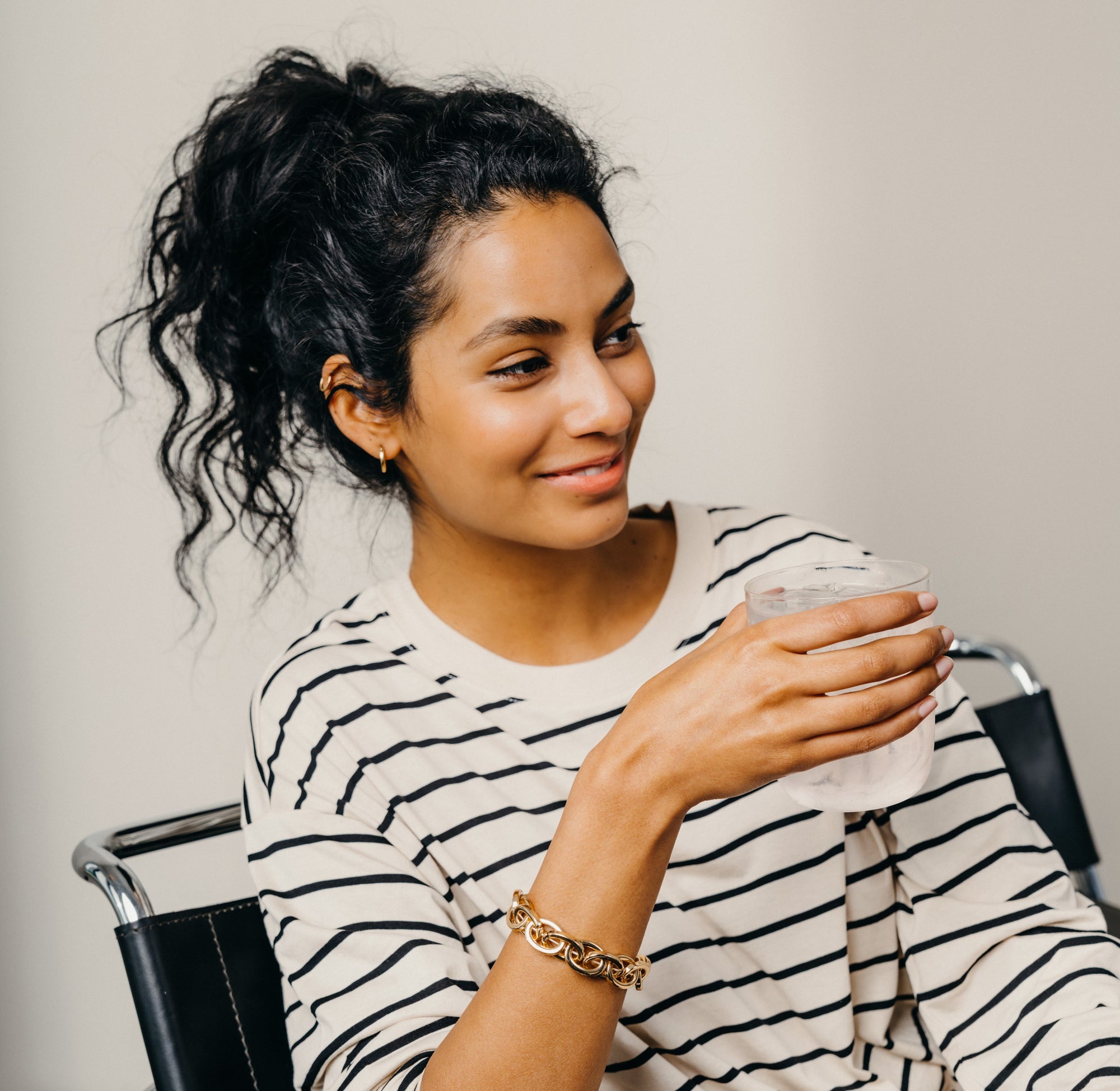 Smiling woman with curly hair wearing a striped shirt, holding a glass of water, showcasing a relaxed and refreshing moment.