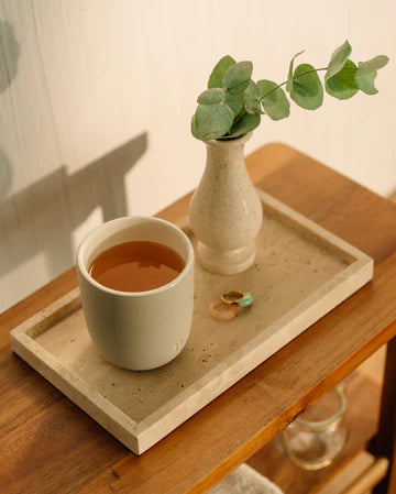 Minimalist tea setup with waterdrop tea cup, eucalyptus vase, and rings on a beige tray on a wooden table in warm sunlight.
