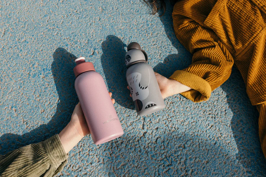 Two hands holding waterdrop bottles against a blue textured background, featuring a pink bottle and a gray animal-themed bottle. Showcasing stylish and sustainable hydration options for all ages.