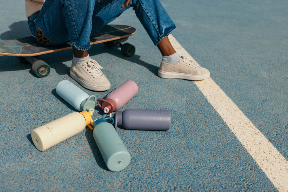 A person sitting on a skateboard wearing casual shoes and jeans, with a selection of colorful waterdrop bottles scattered on the blue pavement beside them.