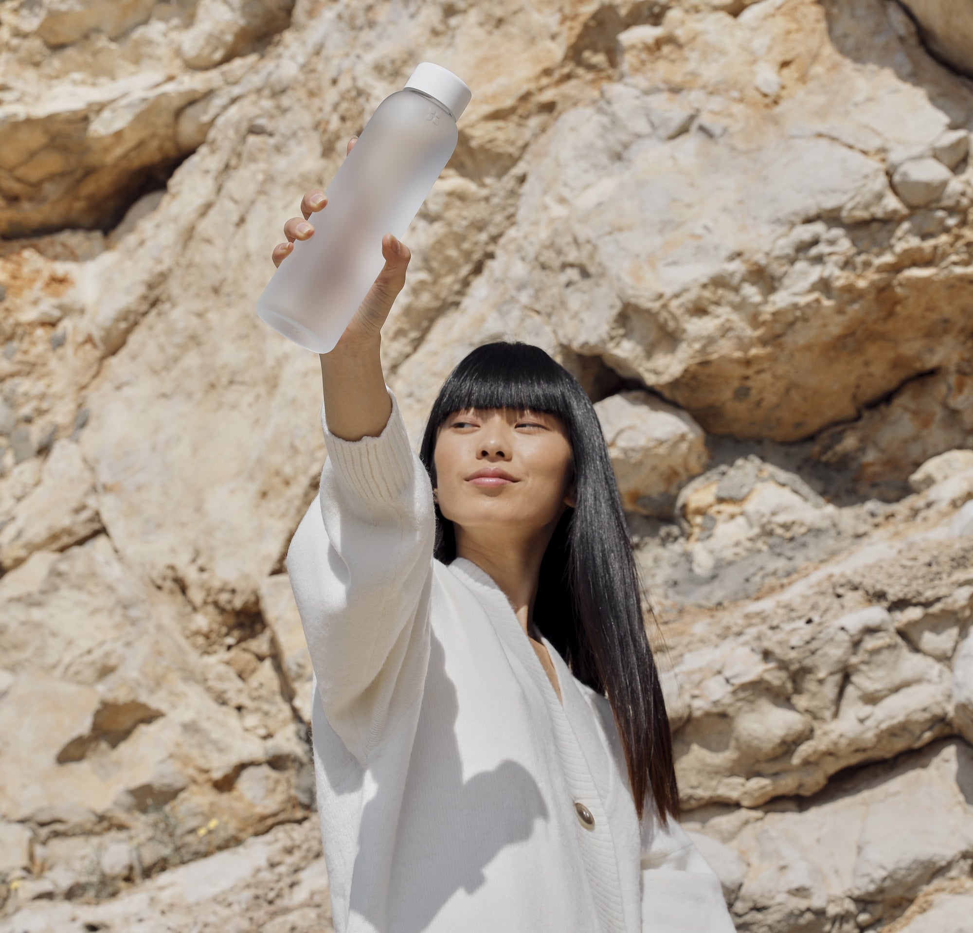 Confident woman holding up a frosted waterdrop bottle against a natural rocky background, embodying a refreshing and sustainable lifestyle.