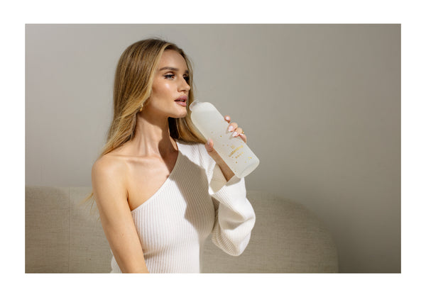 Rosie Huntignton-Whiteley drinking from a frosted waterdrop bottle with gold accents, seated against a neutral background.