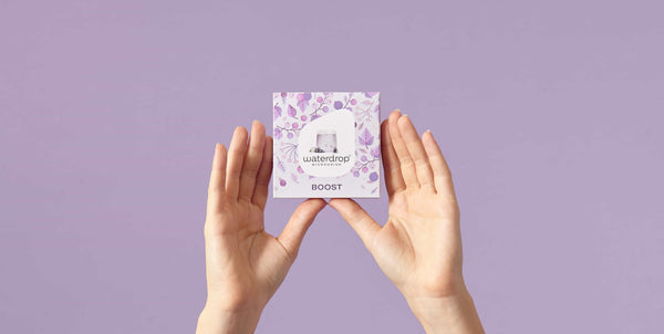 Hands holding Waterdrop BOOST microdrink 12-pack with purple floral design on lavender background.