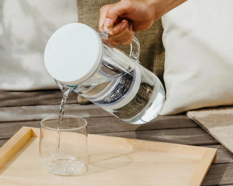 Hand pouring water from a waterdrop glass pitcher into a glass on a wooden tray, promoting hydration and sustainable drinking practices.
