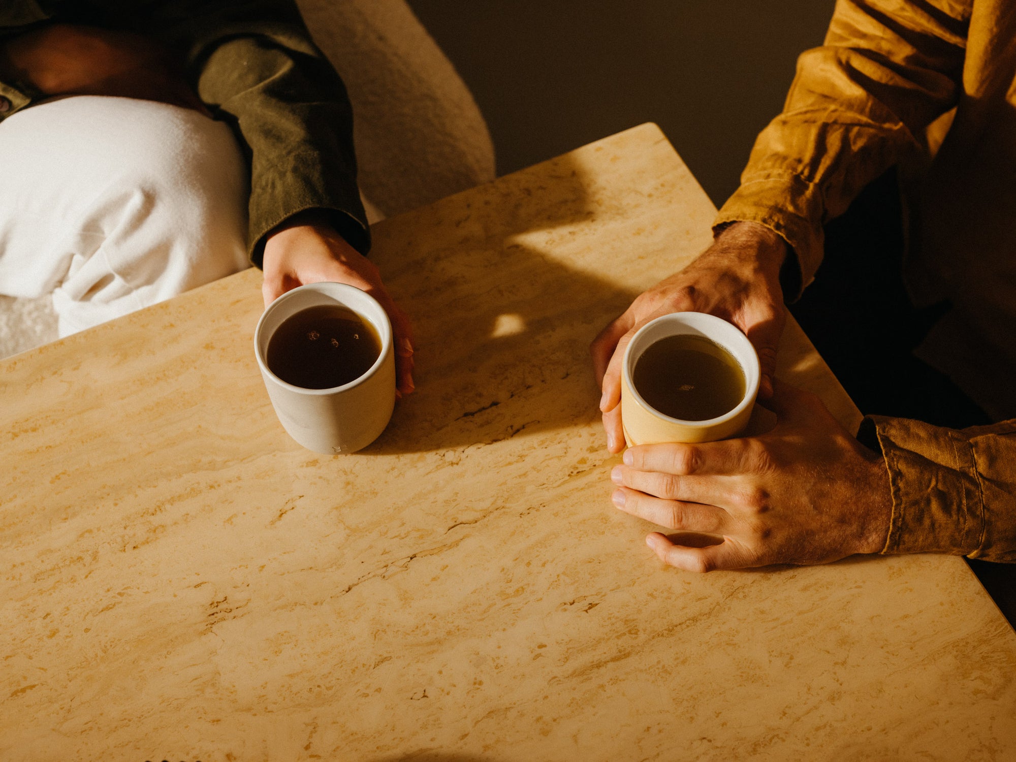 Two people seated at a light-colored table, each holding a cup of waterdrop Microtea, with soft sunlight casting shadows across the scene.