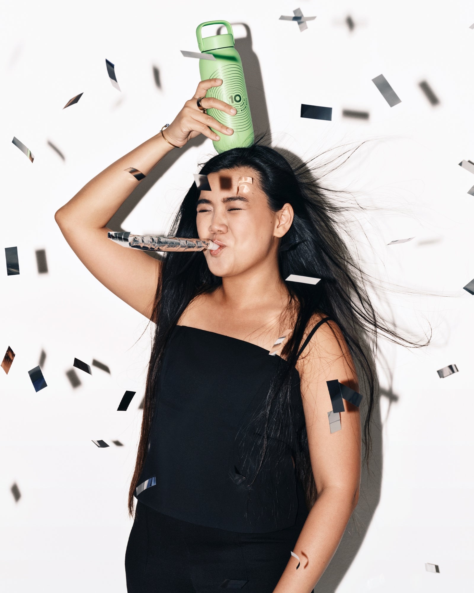 Woman in a black outfit holds a green bottle above her head while confetti falls.
