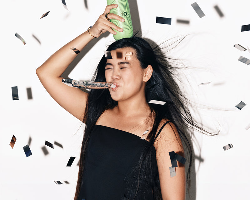 Woman in a black dress celebrates with a party horn and a green spray can, surrounded by confetti.