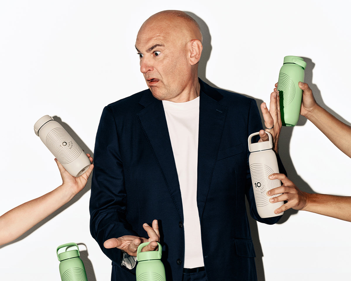 Man in a navy blazer and white shirt surrounded by hands offering several reusable water bottles in different colors.