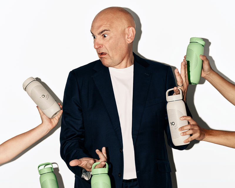 Man in a navy blazer and white shirt surrounded by hands offering several reusable water bottles in different colors.