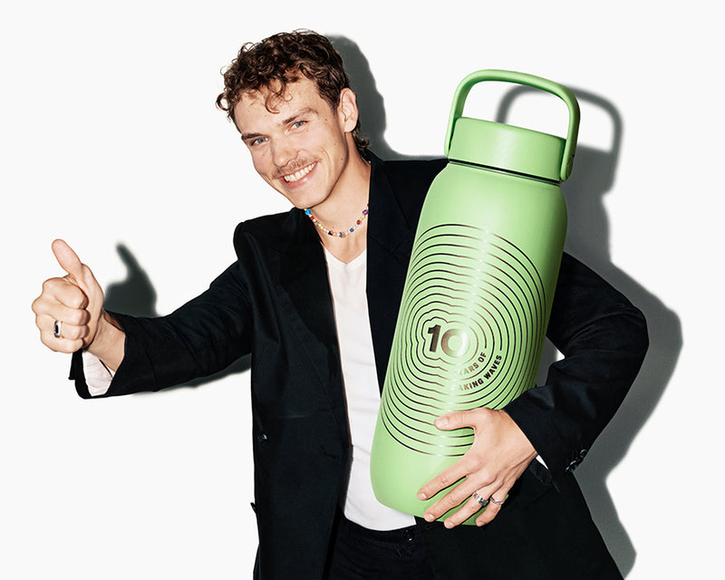 Smiling man in a blazer holds a large green reusable water bottle with a handle and gives a thumbs-up.