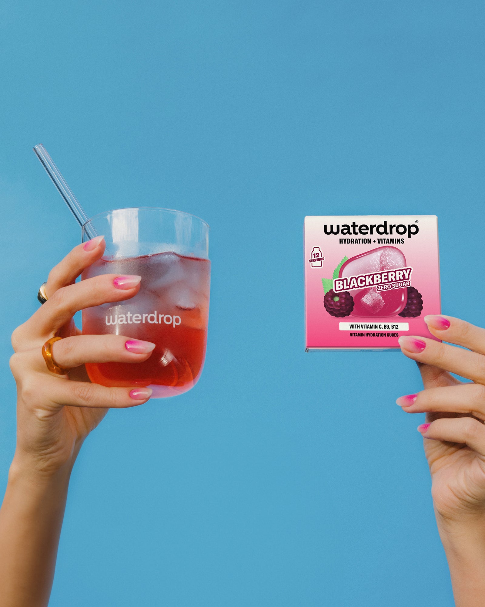 Two hands hold a glass of red juice with a straw and a small pink juice box against a blue background.