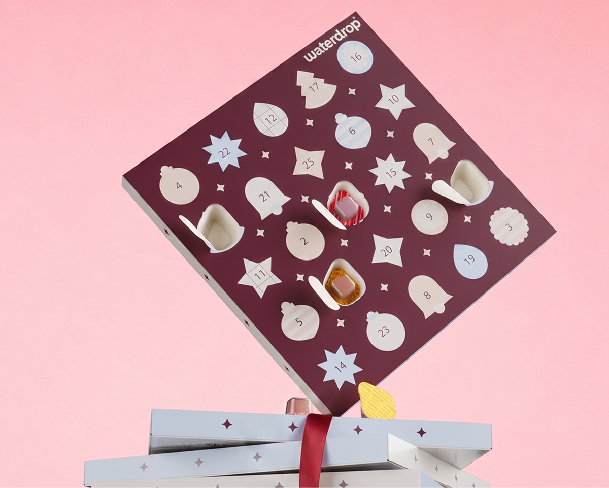 Advent calendar with numbered doors and star-shaped windows revealing treats, placed over gift boxes on a pink background.