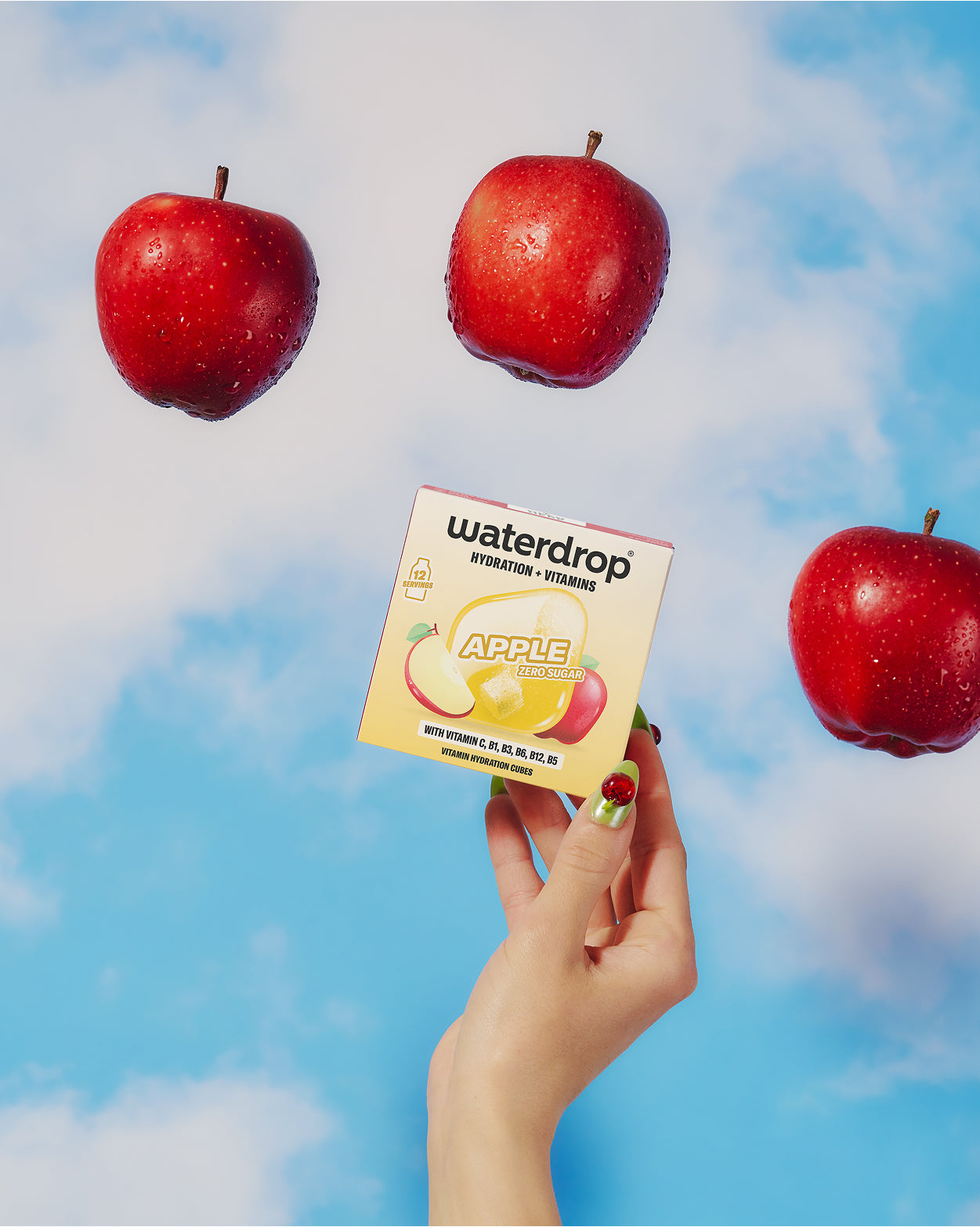 Hand holding a small boxed product with an apple illustration against a blue sky with three floating red apples.