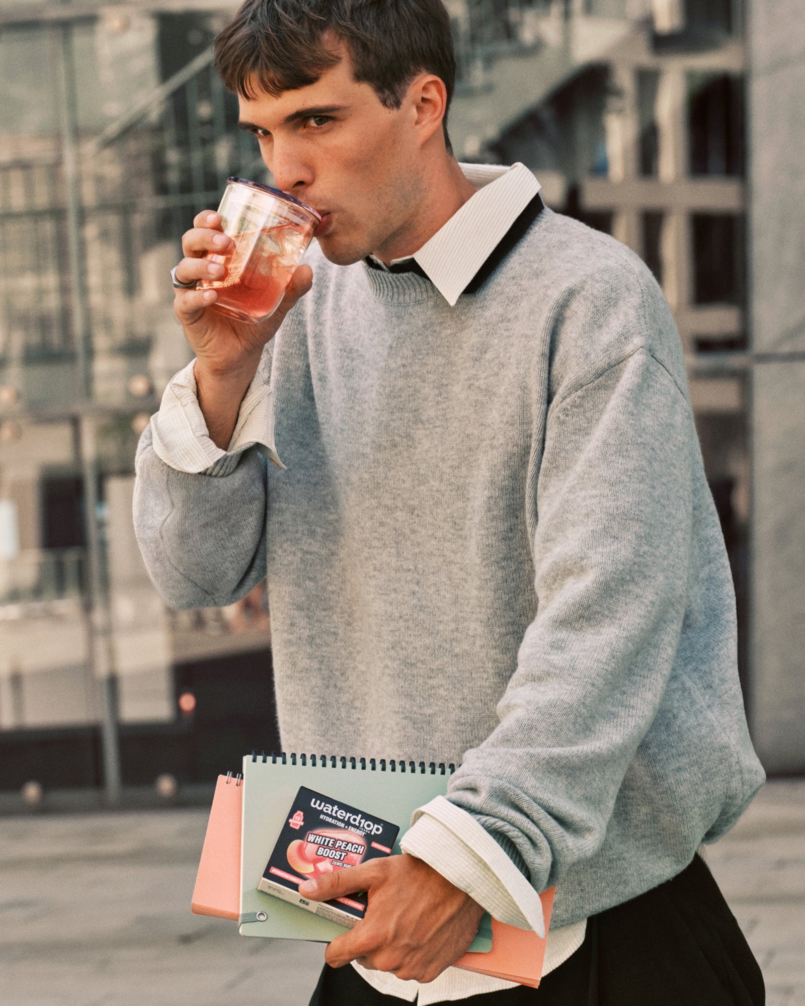 Man in a gray sweater drinks from a cup while carrying notebooks.