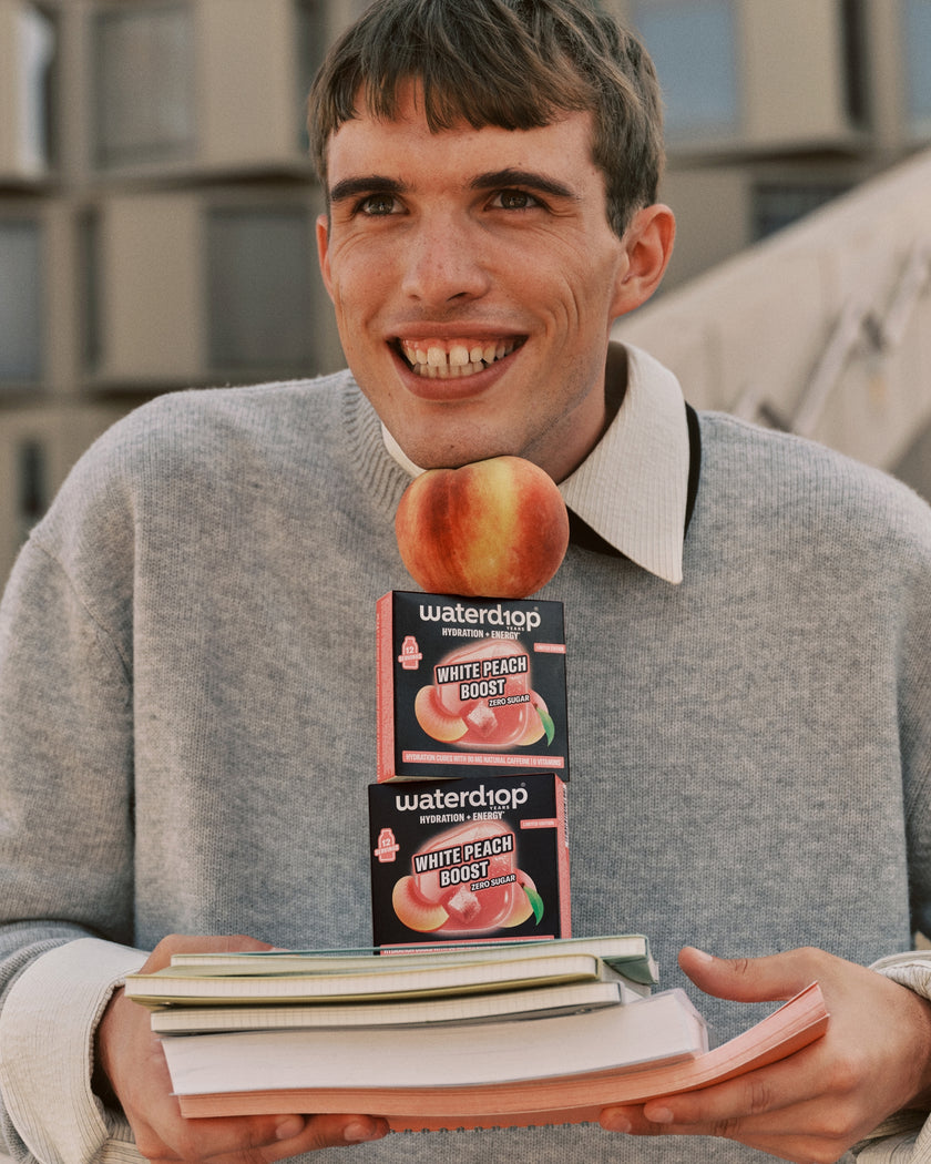 Young man in a gray sweater holding two peach-flavored boxes atop a stack of books.