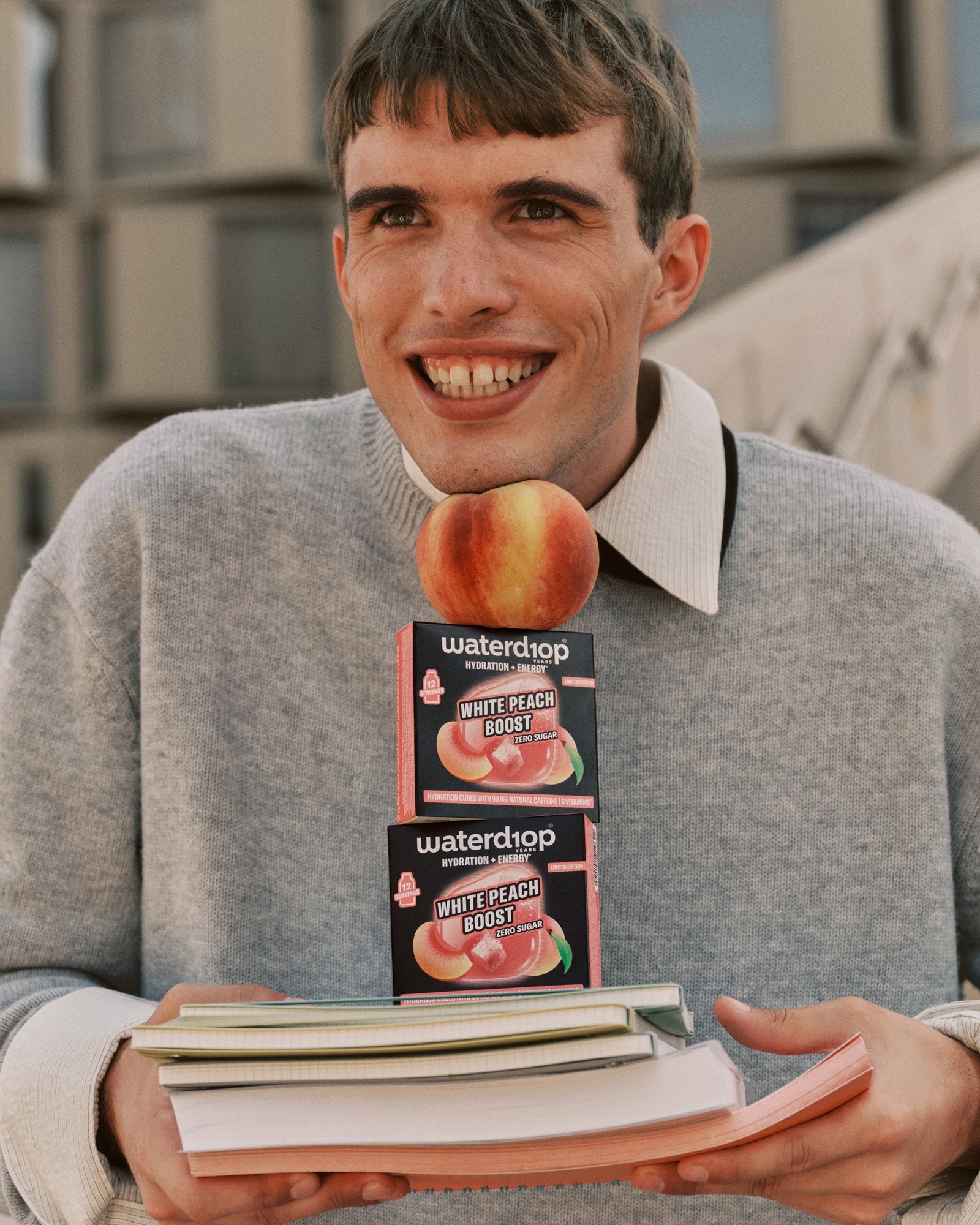 Young man in a gray sweater holding two peach-flavored boxes atop a stack of books.