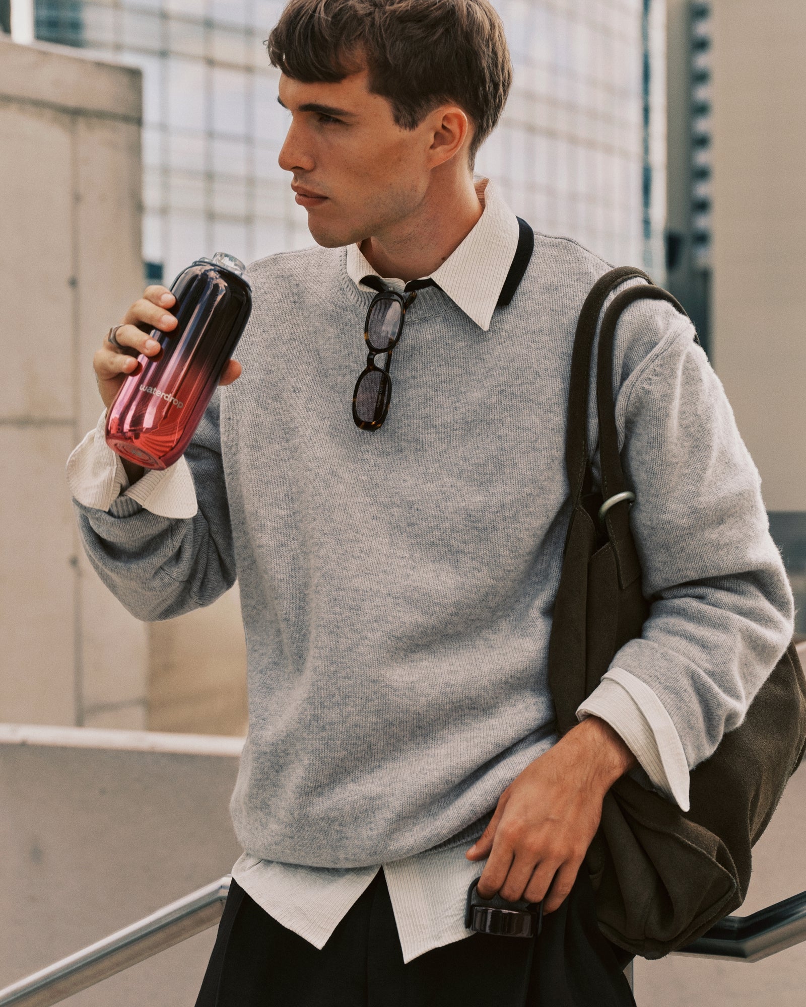 Young man in a gray sweatshirt over a white shirt carries a backpack while drinking from a gradient tumbler.