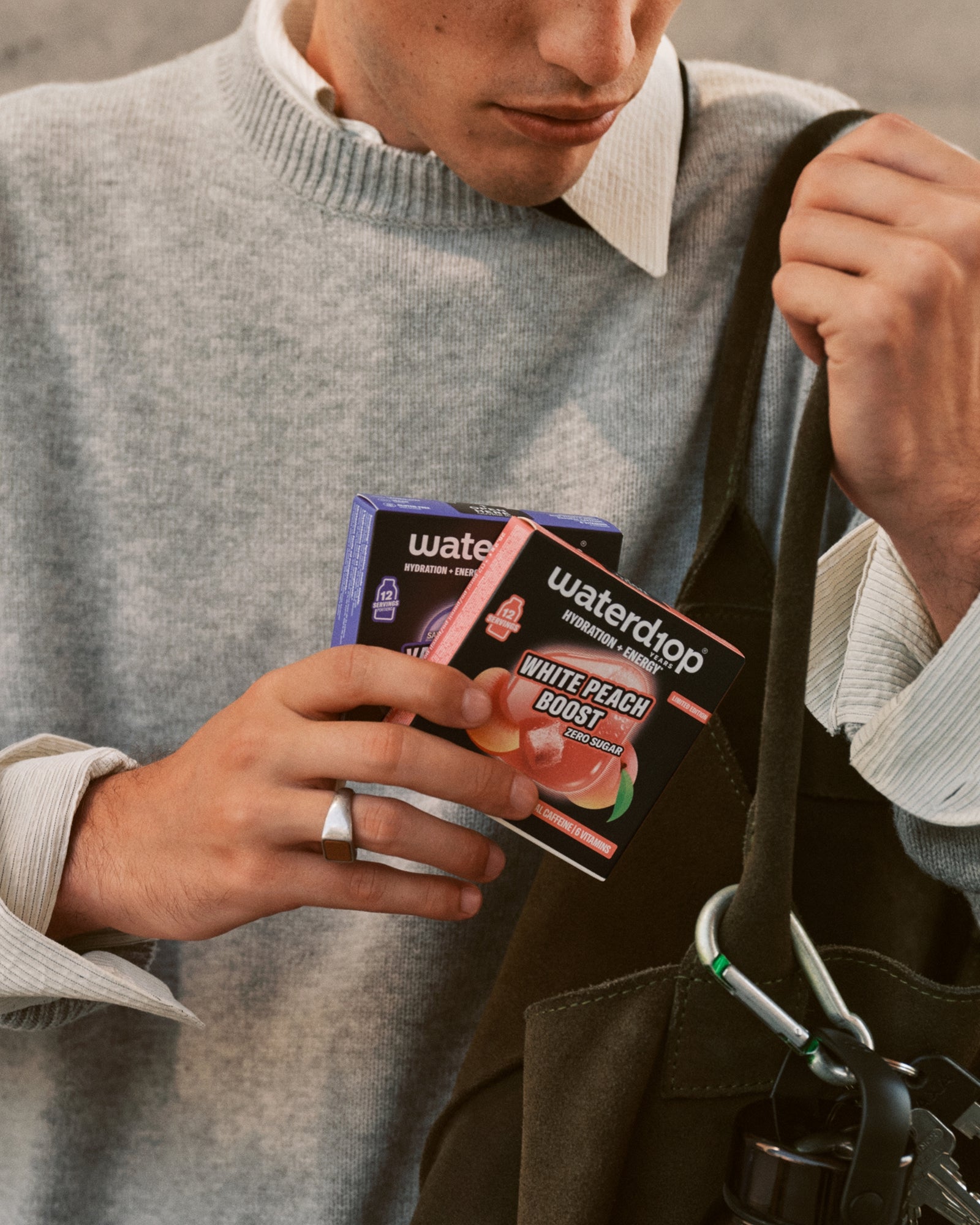 Person in a gray sweater holding small product boxes beside a black tote bag.