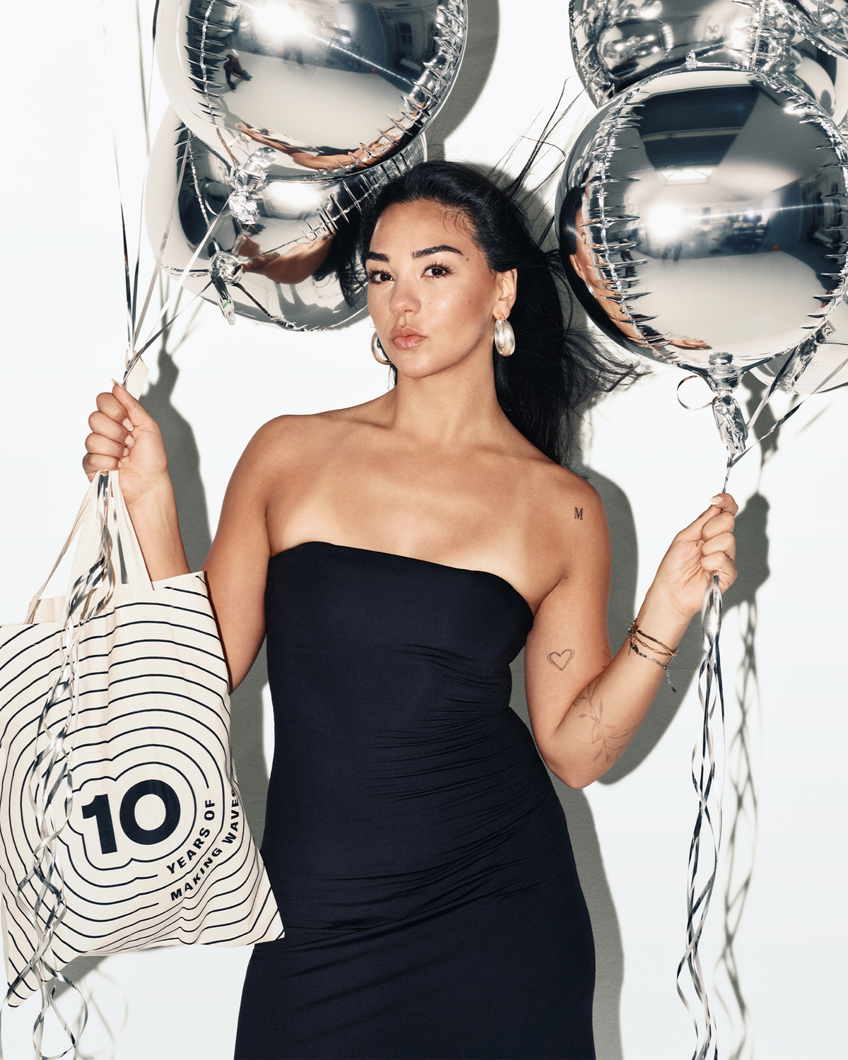 Woman in a strapless black dress holding silver balloons and a striped tote bag.