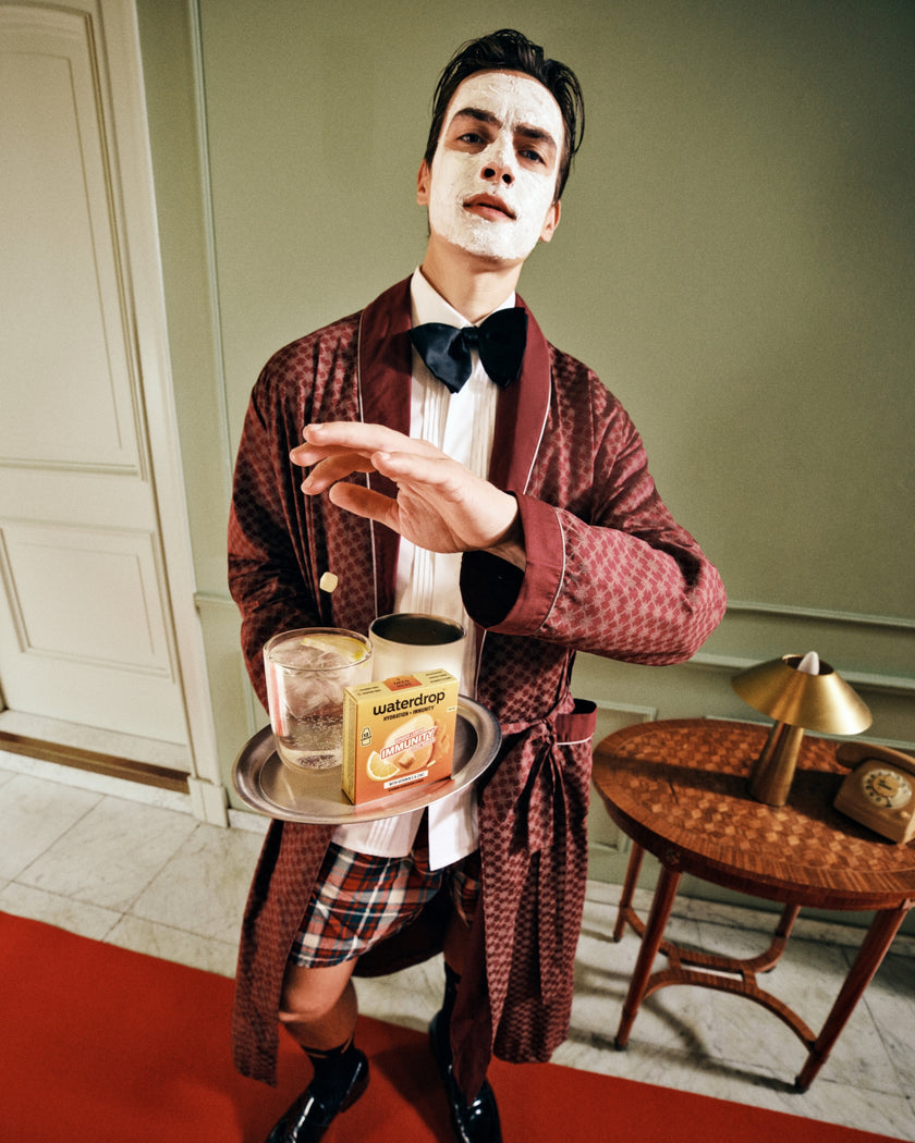 Person wearing white face makeup, burgundy robe and bow tie holds a tray with two glasses.