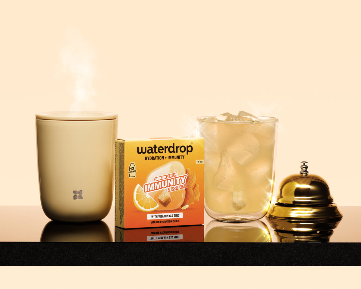 Steaming beige mug, boxed immunity product, glass with ice, and a polished gold bell on a black countertop.