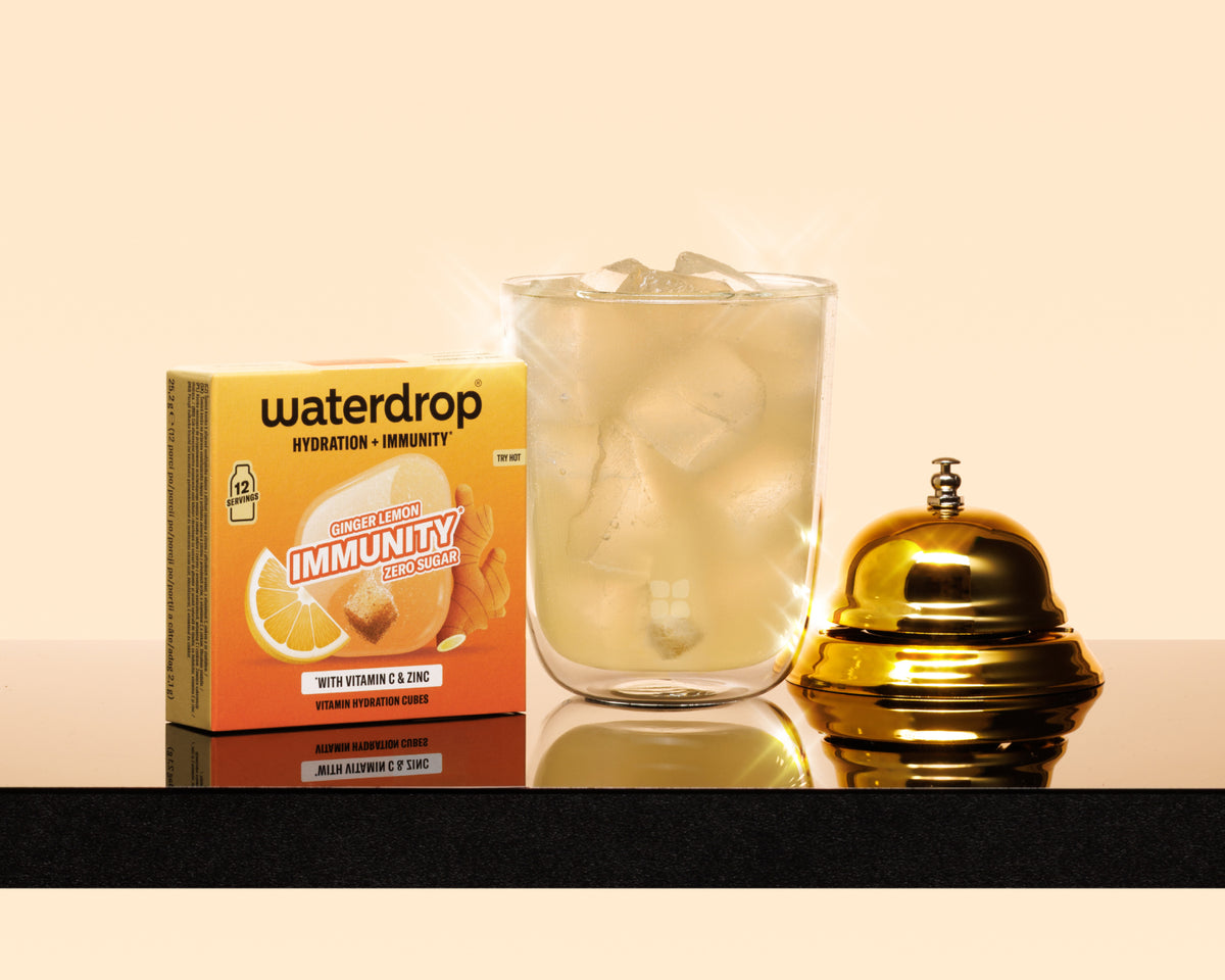 Boxed hydration and immunity product, a glass with ice, and a gold service bell on a dark countertop.