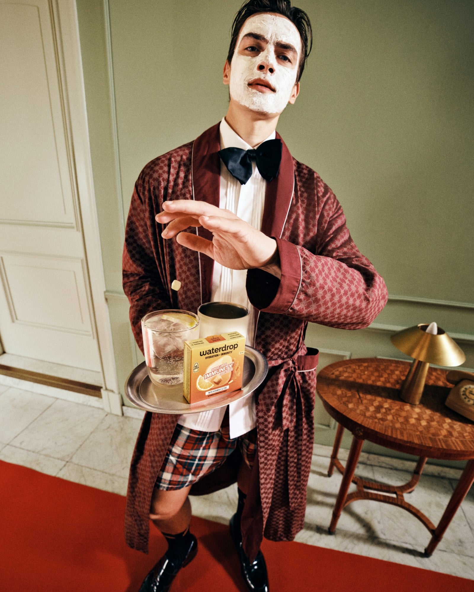 Mime dressed in a burgundy robe and bow tie holds a metal tray with a bottle and glass, standing on a red rug in a vintage-style room.